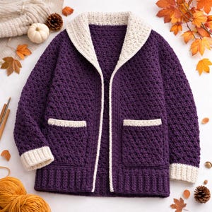May include: A handmade, purple crochet cardigan with a cream shawl collar, pocket trim, and cuffs. The open-front sweater features two front pockets and a textured stitch pattern. The cardigan is displayed on a white surface with autumn decorations.