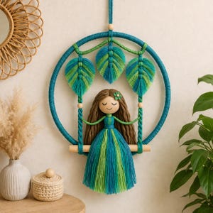 May include: A handmade macrame wall hanging featuring a doll on a swing. The doll has long brown hair and a green and blue dress. The swing is suspended within a teal hoop with green and blue leaf accents. A decorative piece for a nursery or bedroom.