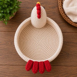 Puede incluir: Una cesta de ganchillo hecha a mano, de color crema, con forma de gallina. La cesta presenta una cabeza de gallina con cresta roja y detalles amarillos, y patas rojas. La cesta est&aacute; sobre una superficie de madera.