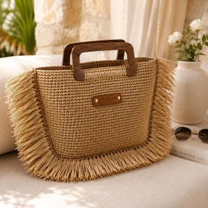 May include: A woven straw tote bag with a rectangular shape and wooden handles. The bag features a fringe trim and a small leather tag that reads "HANDMADE CROCHET". The bag is tan in color and appears to be handmade.