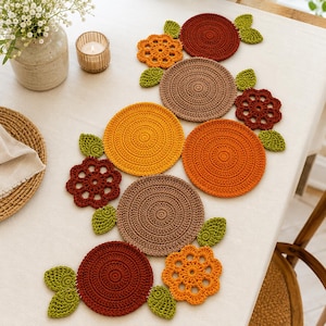 May include: A collection of handmade crochet table decorations. The set includes circular coasters in shades of orange, brown, and yellow, along with flower and leaf accents. The items are arranged on a white tablecloth, creating a warm, inviting aesthetic.