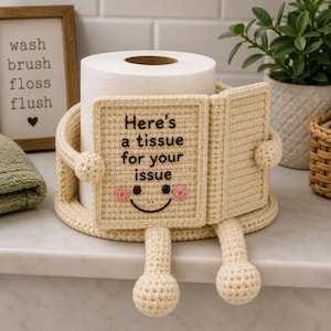 May include: A cream-colored crocheted toilet paper holder shaped like a book, with arms and legs. The book cover reads "Here's a tissue for your issue" with a smiling face. A roll of toilet paper sits inside the holder.
