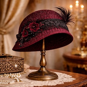 Può includere: Un cappello cloche bordeaux con una fascia in pizzo nero e un'applicazione di piume. Il cappello è decorato con rose e perline bordeaux. È esposto su un espositore dorato, mettendo in risalto il suo stile vintage e i dettagli intricati.