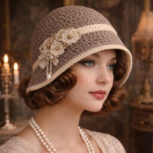 Puede incluir: Un sombrero cloche de ganchillo marrón con una banda y un borde color crema. El sombrero está adornado con flores de ganchillo color crema, perlas y un lazo. El sombrero tiene un estilo vintage de la época de los años 20.