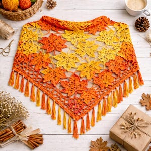 Puede incluir: Un chal de ganchillo hecho a mano en colores otoñales naranja y amarillo, con un diseño de hojas. El chal tiene forma triangular con flecos en el borde inferior. Se muestra sobre una superficie de madera blanca.