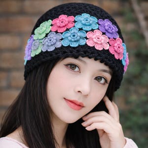 Peut inclure: Un bonnet noir en crochet orné de fleurs colorées en trois dimensions, dans les tons de rose, bleu, violet et vert. Le chapeau est porté sur la tête d'une personne, avec les fleurs disposées autour du bord. Un accessoire élégant.