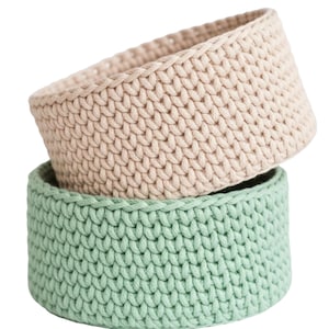 May include: Two handmade crochet baskets. The top basket is a light beige color, and the bottom basket is a soft green. The baskets are round and have a textured, woven appearance. They are stacked on top of each other.