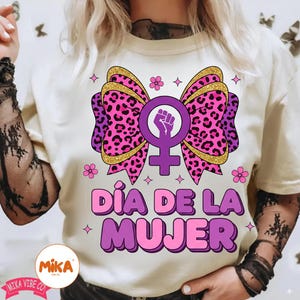 May include: A cream-colored t-shirt featuring a large graphic of a pink and purple leopard print bow centered behind a purple female gender symbol with a raised fist. The text D&Iacute;A DE LA MUJER is printed in bold purple letters below the bow.