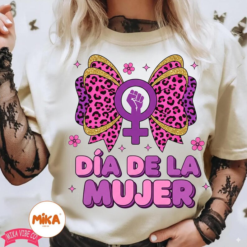 May include: A cream-colored t-shirt featuring a large graphic of a pink and purple leopard print bow centered behind a purple female gender symbol with a raised fist. The text D&Iacute;A DE LA MUJER is printed in bold purple letters below the bow.