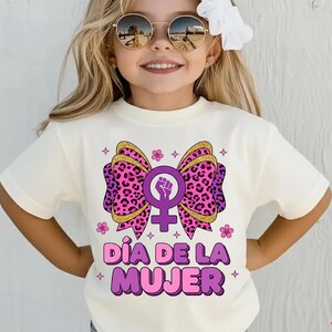 May include: Cream-colored t-shirt featuring a large bow design with a pink leopard print, gold glitter accents, and a purple feminist symbol. The text "D&Iacute;A DE LA MUJER" is displayed in purple. The shirt is for girls.