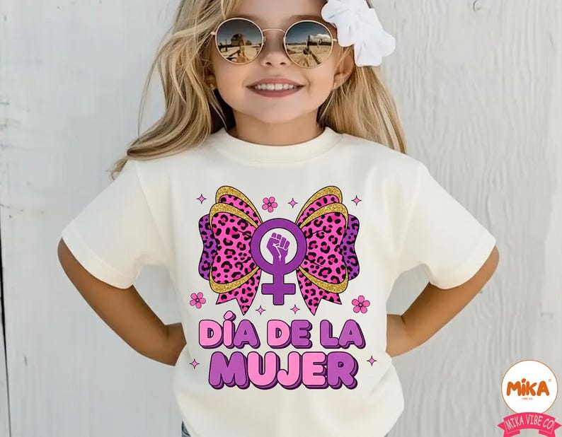 May include: Cream-colored t-shirt featuring a large bow design with a pink leopard print, gold glitter accents, and a purple feminist symbol. The text "D&Iacute;A DE LA MUJER" is displayed in purple. The shirt is for girls.