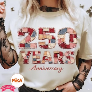 Puede incluir: Camiseta color crema con el texto "250 YEARS Anniversary" en un diseño de patchwork. Los números y letras están compuestos por cuadrados de tela en tonos rojos, azules y beige. La camiseta tiene cuello redondo.