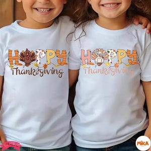 May include: Two white t-shirts with a Thanksgiving theme. Each shirt features the word "HAPPY" in orange and yellow block letters, with a turkey illustration and the words "Thanksgiving" in brown script. The shirts are worn by two children.
