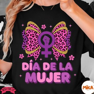 May include: Black t-shirt featuring a pink and gold bow with a leopard print design, a purple female symbol with a raised fist, and the text "D&iacute;a de la Mujer" in pink and purple. The shirt has a feminine theme.