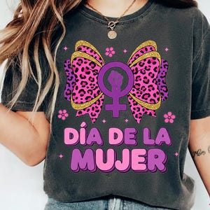 May include: Dark gray t-shirt featuring a graphic design for International Women's Day. The design includes a purple bow with a leopard print, a female symbol with a raised fist, and the text "D&iacute;a de la Mujer" in pink and purple.
