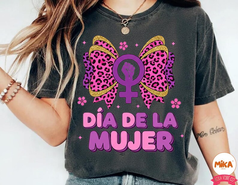 May include: Dark gray t-shirt featuring a graphic design for International Women's Day. The design includes a purple bow with a leopard print, a female symbol with a raised fist, and the text "D&iacute;a de la Mujer" in pink and purple.
