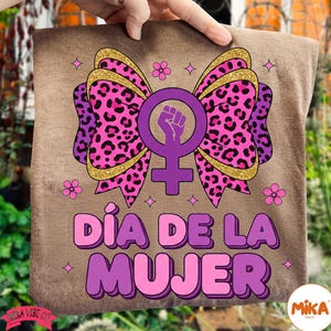 May include: A brown t-shirt featuring a large bow with a leopard print design in pink and purple, accented with gold glitter. The bow surrounds a purple female symbol with a raised fist. The text "D&Iacute;A DE LA MUJER" is displayed in purple and pink.