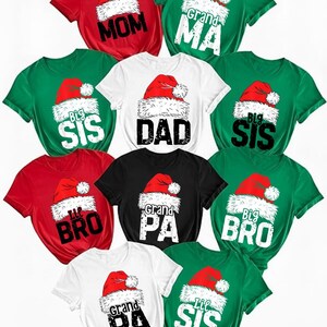 May include: A collection of Christmas-themed t-shirts in red, green, black, and white. Each shirt features a Santa hat graphic and text such as "Mom," "Dad," "Big Sis," and "Grandpa." The shirts are short-sleeved.