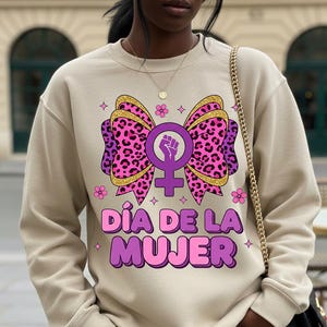 May include: Beige sweatshirt featuring a large graphic of a pink leopard print bow with a gold glitter outline, a purple female symbol with a raised fist, and the text "D&iacute;a de la Mujer" in pink and purple. Small pink flowers and stars surround the design.