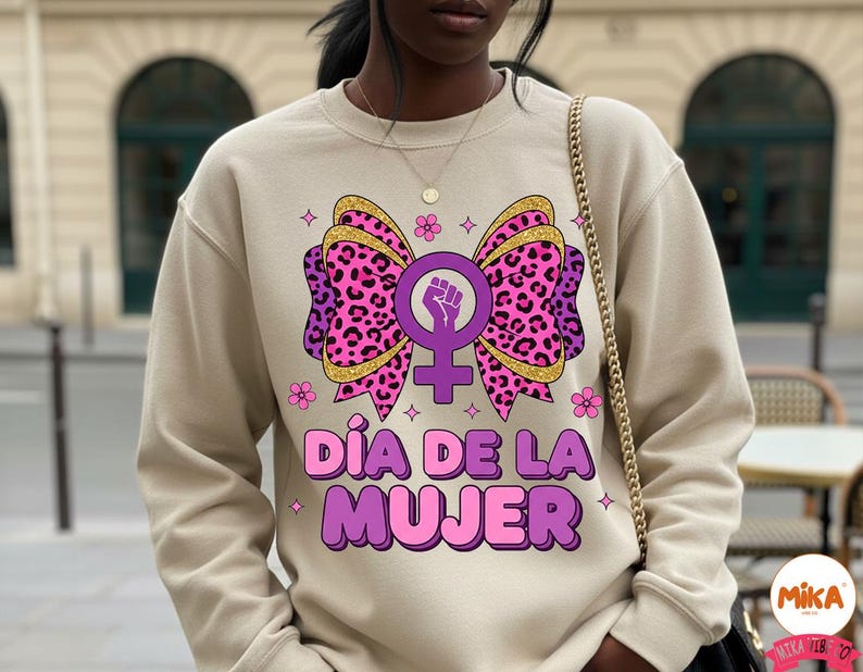 May include: Beige sweatshirt featuring a large graphic of a pink leopard print bow with a gold glitter outline, a purple female symbol with a raised fist, and the text "D&iacute;a de la Mujer" in pink and purple. Small pink flowers and stars surround the design.