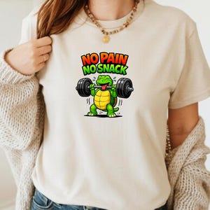 May include: A beige t-shirt featuring a cartoon turtle lifting weights. The turtle is green with a yellow shell. The text "NO PAIN NO SNACK" is above the turtle in red and green letters.