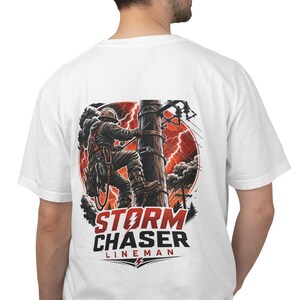 May include: A white t-shirt featuring a graphic on the back of a utility worker climbing a wooden pole during a storm with lightning and dark clouds. The text below the illustration reads STORM CHASER LINEMAN in bold red and black lettering.
