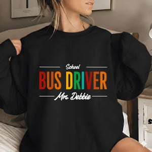 Puede incluir: Sudadera negra con el texto "School BUS DRIVER Mrs. Debbie" en letras coloridas. El texto está centrado en la parte delantera. La sudadera es de cuello redondo.
