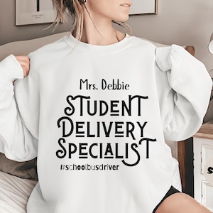 Puede incluir: Sudadera blanca con el texto negro "Mrs. Debbie STUDENT DELIVERY SPECIALIST #schoolbusdriver". La sudadera es de cuello redondo y parece estar hecha de un material suave. El texto está centrado en la parte delantera.
