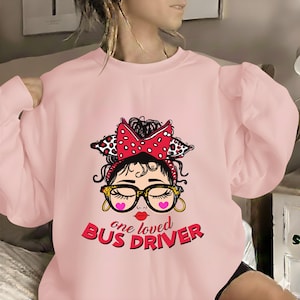 Puede incluir: Sudadera rosa claro con un gráfico de una mujer con gafas, un lazo rojo y el texto "one loved BUS DRIVER". El diseño incluye corazones y un labio rojo. La sudadera es de cuello redondo.