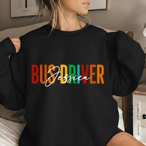 Puede incluir: Sudadera negra con el texto "BUS DRIVER Jessica" en letras de bloque coloridas. La palabra "BUS" es roja, "DRIVER" es verde y naranja, y "Jessica" está en una fuente cursiva blanca.