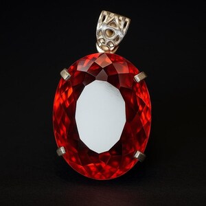 Può includere: Un ciondolo con pietra preziosa rossa di forma ovale e sfaccettata, montato su una montatura in metallo argentato con griffe. Il ciondolo presenta una contromaglia decorativa in metallo traforato sulla parte superiore, con un riflesso bianco.