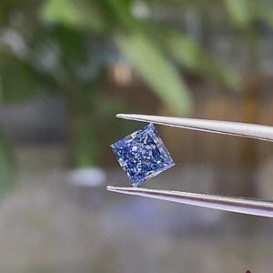 May include: A square-cut, brilliant blue gemstone held by a pair of silver-toned metal tweezers. The stone features multiple facets that reflect light, creating a sparkling appearance with deep and light blue tones throughout the surface of the gem.