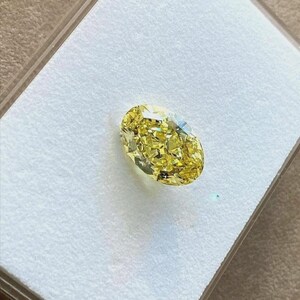 May include: A loose oval-cut gemstone with a vibrant yellow colour and numerous facets that create a brilliant, reflective surface. The stone is displayed resting on a white, textured foam insert inside a clear plastic storage container.