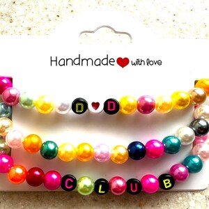 May include: Three beaded bracelets with colorful, round beads. The top bracelet has the letters "D D" with a heart in between. The middle bracelet is a rainbow of colors. The bottom bracelet spells out "CLUB". The card reads "Handmade with love".