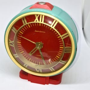 Può includere: Una sveglia vintage con cassa turchese, quadrante rosso e numeri romani dorati. L'orologio ha una base e una parte superiore rosse. Il quadrante recita "Jantar" e "4 Jewels Made in UK".