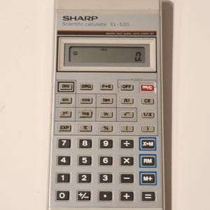 Peut inclure: Une calculatrice scientifique SHARP EL-530 vintage. La calculatrice est gris clair avec des boutons noirs et bleus. L'écran affiche le chiffre 0. La calculatrice possède diverses touches de fonction, dont des fonctions trigonométriques et de mémoire.