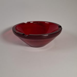 Peut inclure: Un cendrier en verre rouge profond avec une forme de bol circulaire lisse. Le bord présente deux encoches distinctes conçues pour poser des objets. Le verre est translucide, laissant passer la lumière à travers le matériau rouge vif.