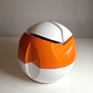 Può includere: Oggetto sferico bicolore, con parte superiore e base bianche, e sezione centrale arancione vivace. Una sottile striscia grigio argento adorna la parte superiore. Probabilmente un oggetto decorativo o un contenitore, dal design moderno.