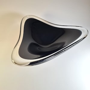 Op de afbeelding: Een moderne, driehoekige kom met een glanzend zwart interieur en een heldere, afgeronde rand. Het ontwerp van de kom is strak en minimalistisch, reflecteert licht en creëert een gevoel van diepte. De kom is waarschijnlijk gemaakt van glas of een vergelijkbaar materiaal.
