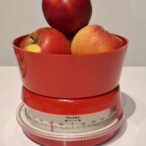 Pode incluir: Uma balança de cozinha vermelha vintage com uma tigela cheia de maçãs vermelhas. A balança tem um mostrador branco com números pretos e a marca "POLONA". As maçãs são uma mistura de vermelho e amarelo.