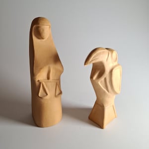 May include: Two tan-colored, carved wooden sculptures. One is a tall, stylized figure with a head covering and scales of justice. The other is a stylized bird-like figure. Both are on a white background.