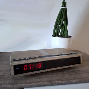 Op de afbeelding: Een zilverkleurige digitale wekker met de tijd 07:18 in rode cijfers. De wekker heeft knoppen en een luidsprekerrooster. Achter de wekker staat een witte pot met groene, cilindervormige planten.
