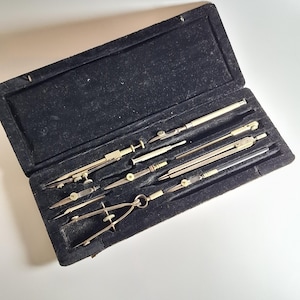 Pode incluir: Uma caixa preta forrada aveludada aberta contendo um conjunto de ferramentas de desenho vintage. O conjunto inclui vários compassos, divisores e outros instrumentos de precisão, todos em prata e ouro.