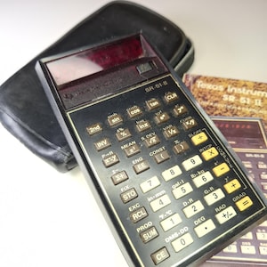 Puede incluir: Una calculadora científica Texas Instruments SR-51-II vintage con una funda negra. La calculadora tiene numerosos botones con funciones matemáticas y una pantalla roja. Se incluye un manual.