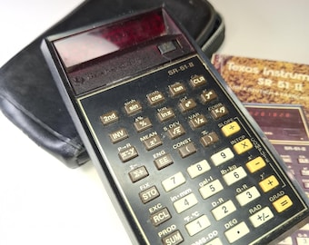 Könnte beinhalten: Ein Vintage Texas Instruments SR-51-II wissenschaftlicher Taschenrechner mit schwarzem Gehäuse. Der Rechner hat zahlreiche Tasten mit mathematischen Funktionen und ein rotes Display. Ein Handbuch ist enthalten.