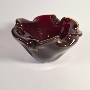 Peut inclure: Un bol en verre bordeaux profond avec un bord ondulé et froncé. Le bol présente une finition lisse et brillante ainsi qu'une qualité translucide qui laisse passer la lumière à travers le matériau rouge foncé, soulignant la forme organique.
