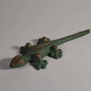 Può includere: Una statuetta di lucertola in metallo fuso con una finitura verde testurizzata e riflessi color rame usurati sulla testa, sul dorso e sulle zampe. La lucertola è raffigurata in posizione strisciante con una coda lunga e sottile verso destra.