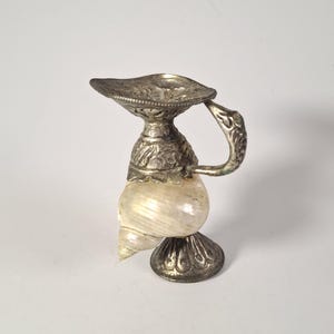 May include: A small decorative pitcher featuring a natural white seashell body accented with ornate silver-toned metal components. The piece includes a detailed metal base, a textured handle, and a wide, embossed metal rim with a pouring spout.