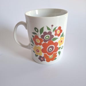 Puede incluir: Una taza de cerámica blanca con un colorido estampado floral en la parte frontal. El diseño consta de flores estilizadas en tonos naranja, rojo, amarillo y morado, acentuadas con hojas verdes. La taza tiene un asa curva en el lado izquierdo.