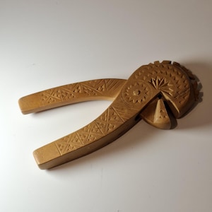Könnte beinhalten: Ein hellbrauner Nussknacker aus Holz mit einem detaillierten Schnitzmuster. Die Griffe und der Kopf des Nussknackers weisen geometrische Muster und ein kreisförmiges Design auf. Der Nussknacker liegt auf einer weißen Oberfläche.